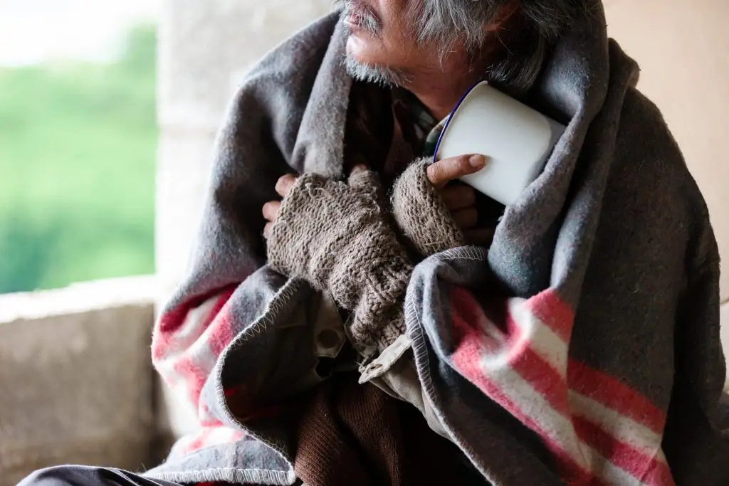 A person with gray hair and beard wrapped in a blanket, holding a white mug while wearing fingerless gloves.