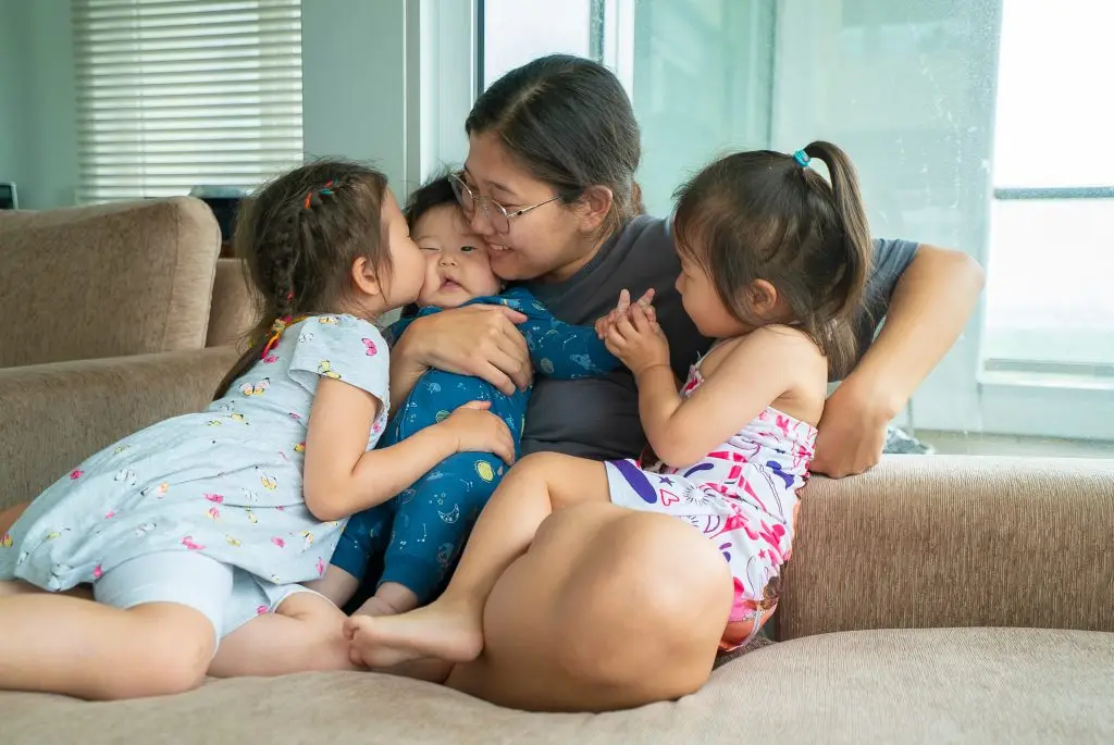 A woman sits on a sofa holding a baby, with a young girl kissing the babys cheek and another girl holding the babys hand.