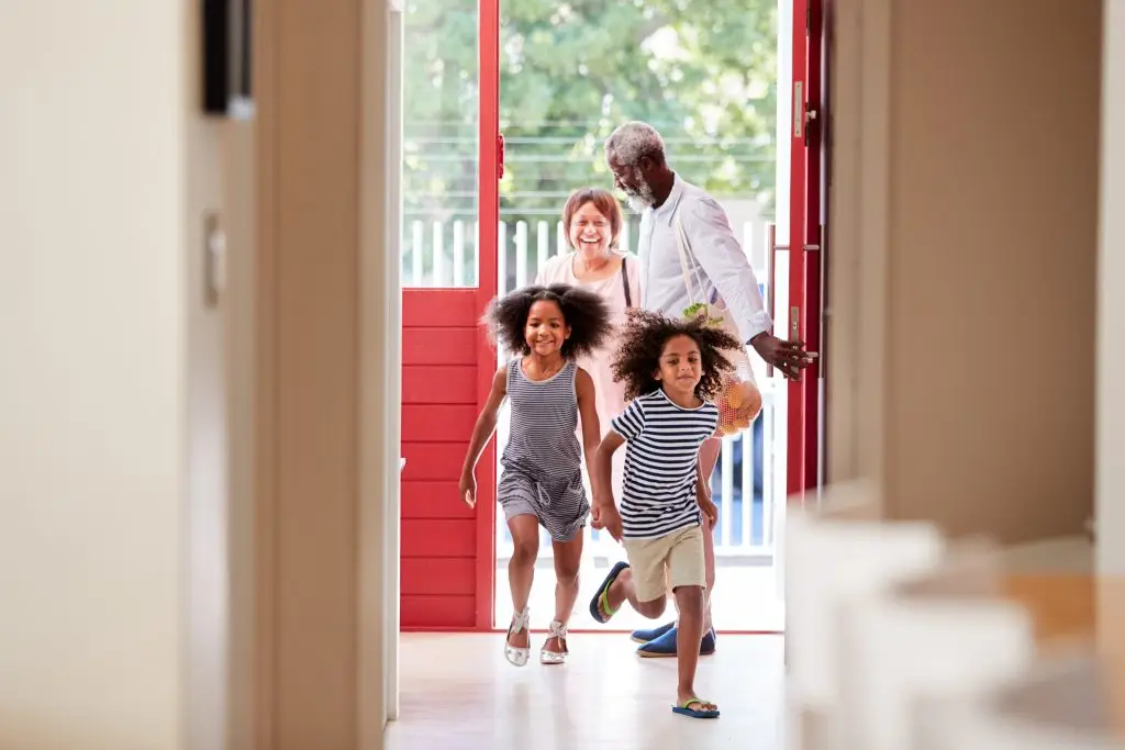 Two children run into a house, followed by their grandparents carrying groceries through an open door.