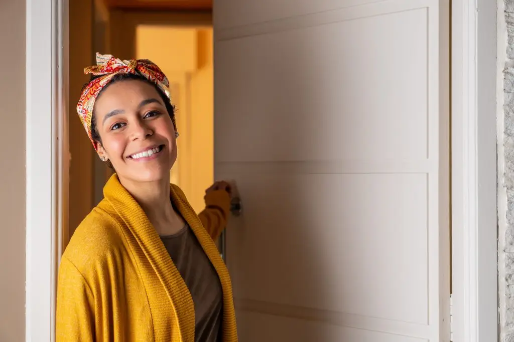 A woman in a yellow cardigan and patterned headscarf smiling while opening a white door.