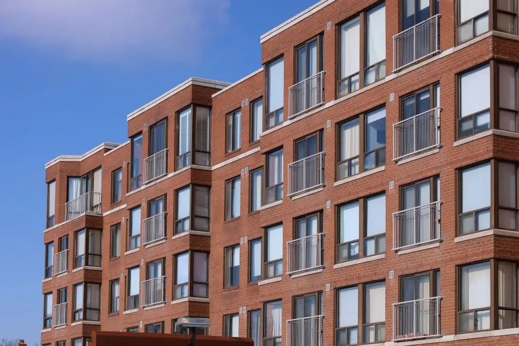 A modern red brick apartment building with rows of balconies and large windows against a blue sky.