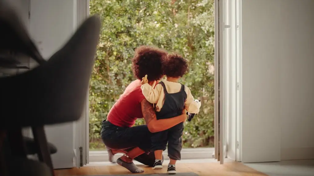 A woman with curly hair kneeling and hugging a toddler in front of an open glass door.
