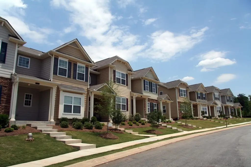A row of identical suburban houses with tan siding and front porches along a paved street.