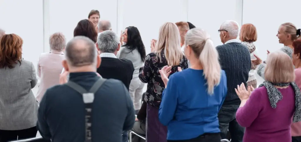 A group of people seen from behind applauding a speaker at the front of a room.